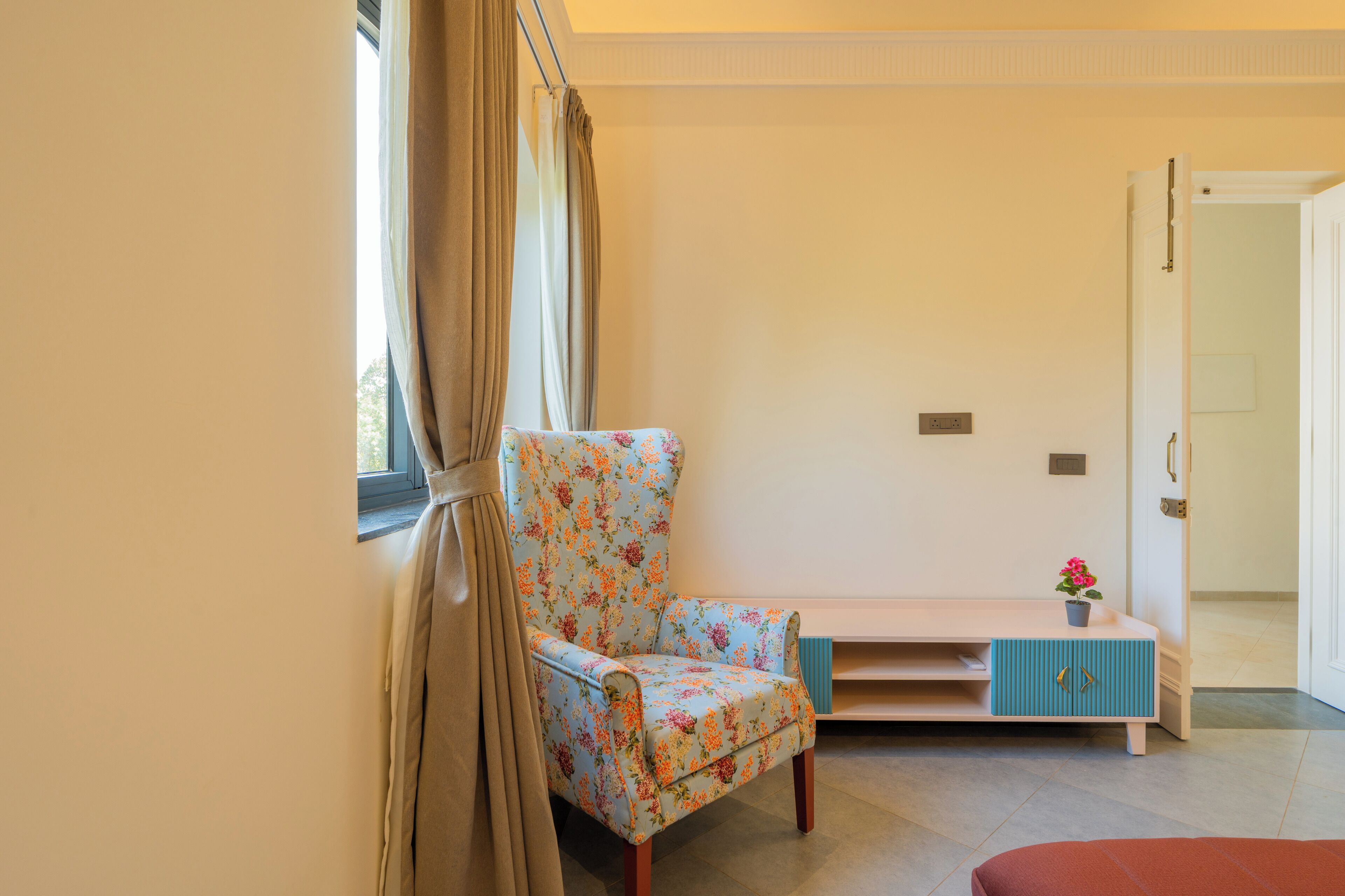 A room with a floral-patterned chair, a small table with a potted plant, and a door leading to another room.