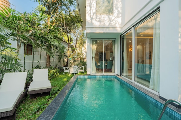 Una piscina con sillas de descanso, una puerta de vidrio que da a una habitación y abundante vegetación.