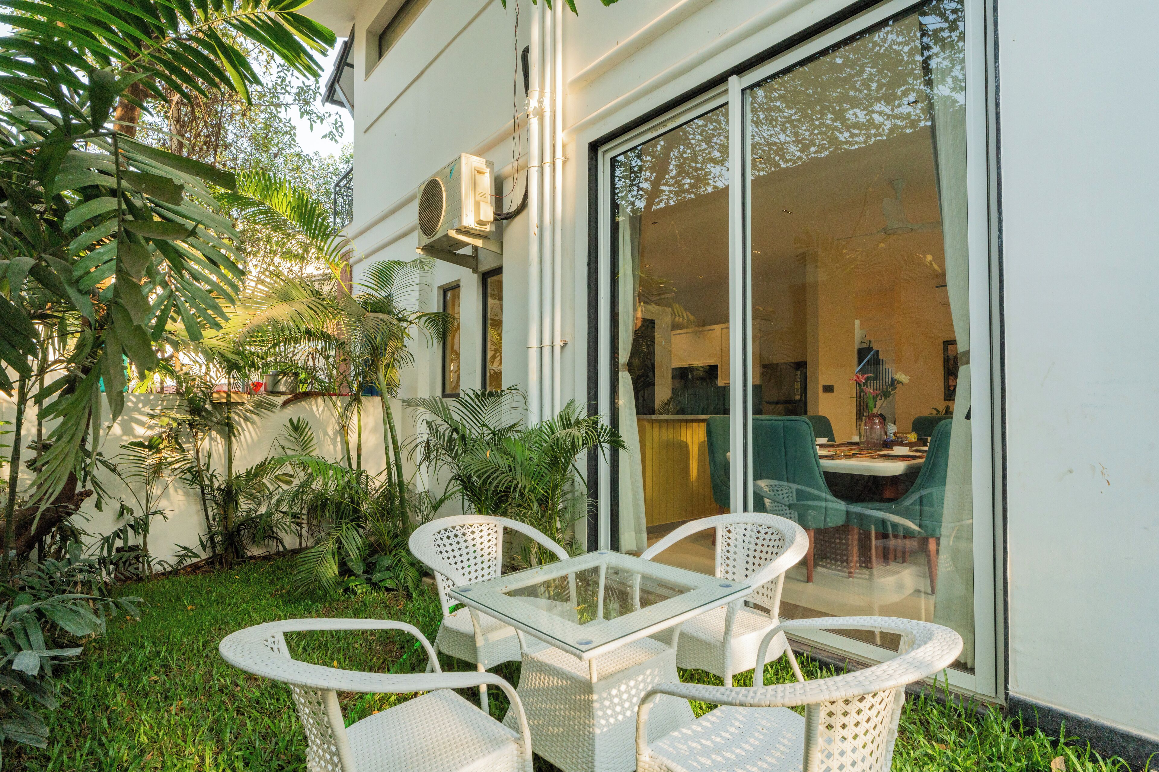 An outdoor patio area with a glass table and white wicker chairs, surrounded by lush greenery and a white building facade.