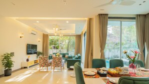 A modern living room with a sofa, dining table, and chairs. There is a TV mounted on the wall, a potted plant, and a window with curtains. - ELIVAAS Laguna Camellia | Luxe 4 BHK Villa Near Popular Beaches with Private Pool (Anjuna)