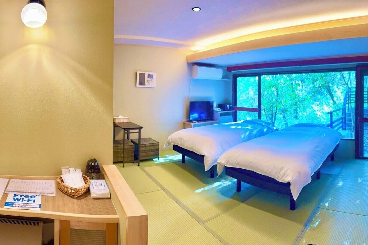 A hotel room with two beds, a desk with a basket, a TV, and a large window with a view of trees.