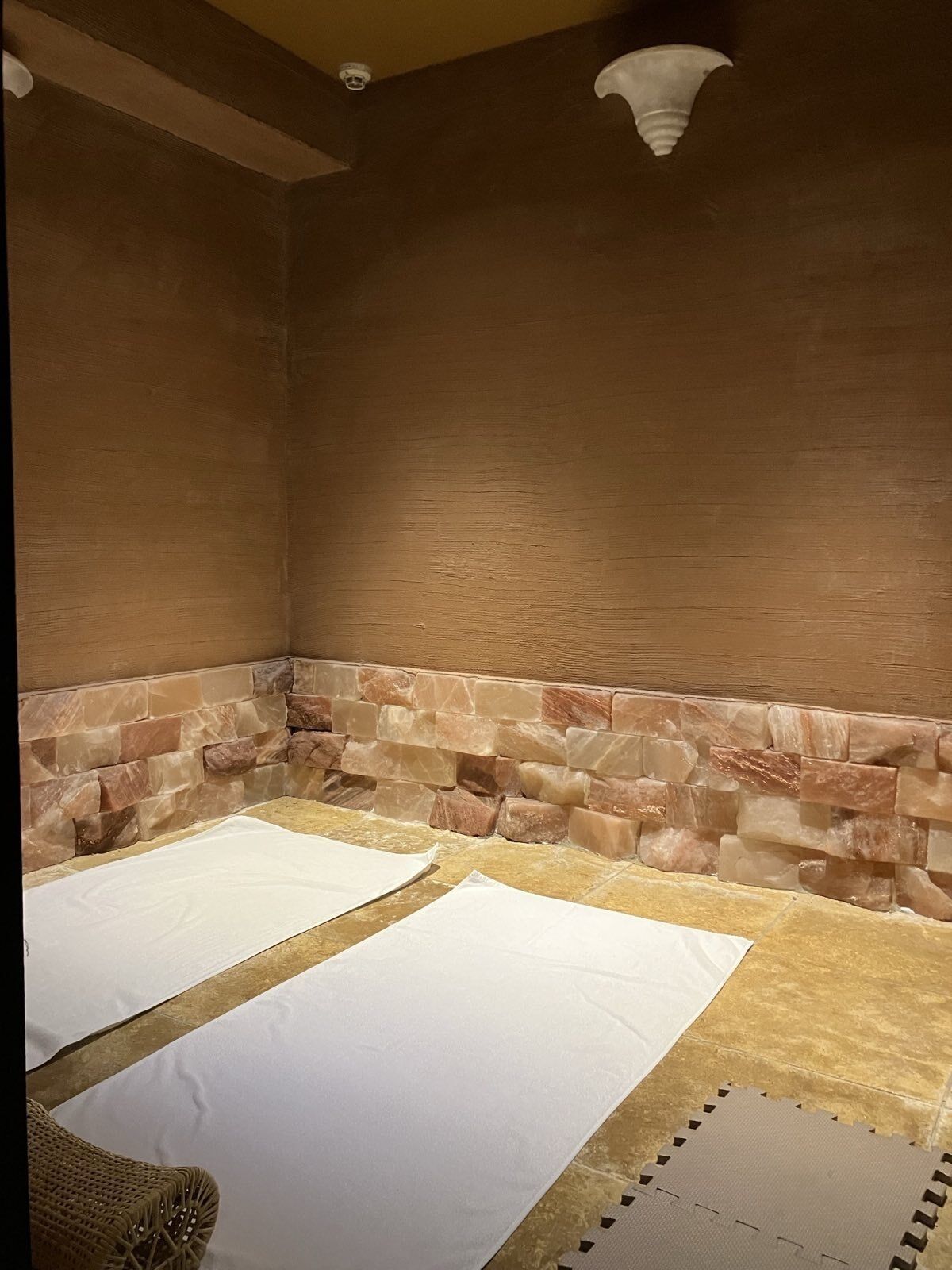 A spa room with a stone wall, two white mats on the floor, and a woven chair.
