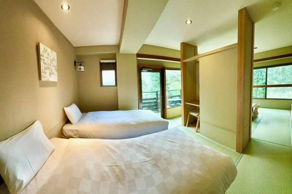 A room with two beds, a large window, and a view of greenery outside.