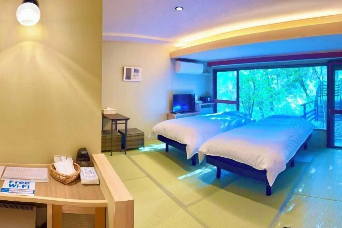A hotel room with two beds, a desk with a basket, a TV, and a large window with a view of trees.