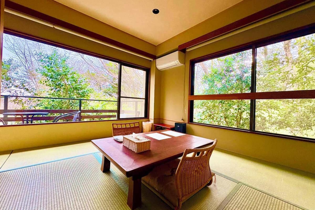 A traditional Japanese room with tatami flooring, a wooden table, and large windows offering a view of trees.