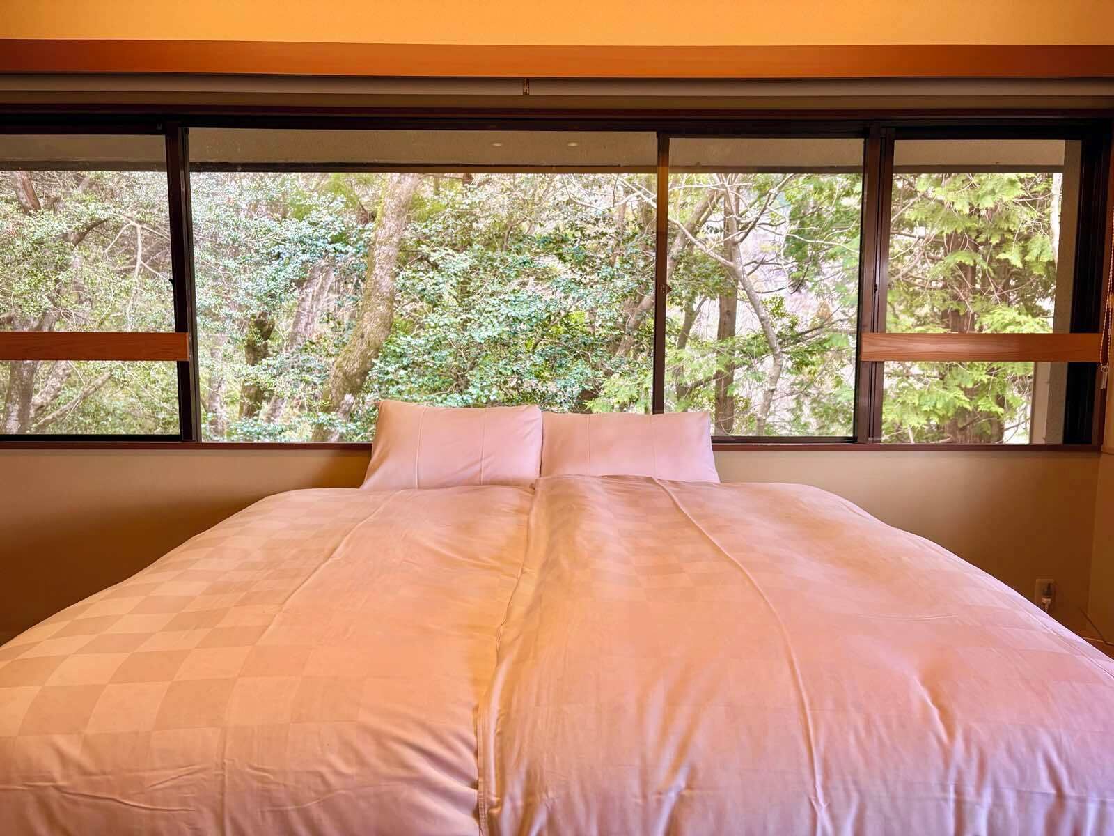 Ein Schlafzimmer mit einem großen Bett, Blick auf Bäume durch das Fenster und einem hölzernen Kopfteil.