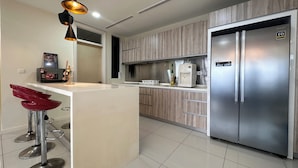 A modern kitchen with a stainless steel refrigerator, wooden cabinets, a bar stool area, and a pendant light. - Skyline Luxe 4BR Retreat with En-suites (Petaling Jaya)