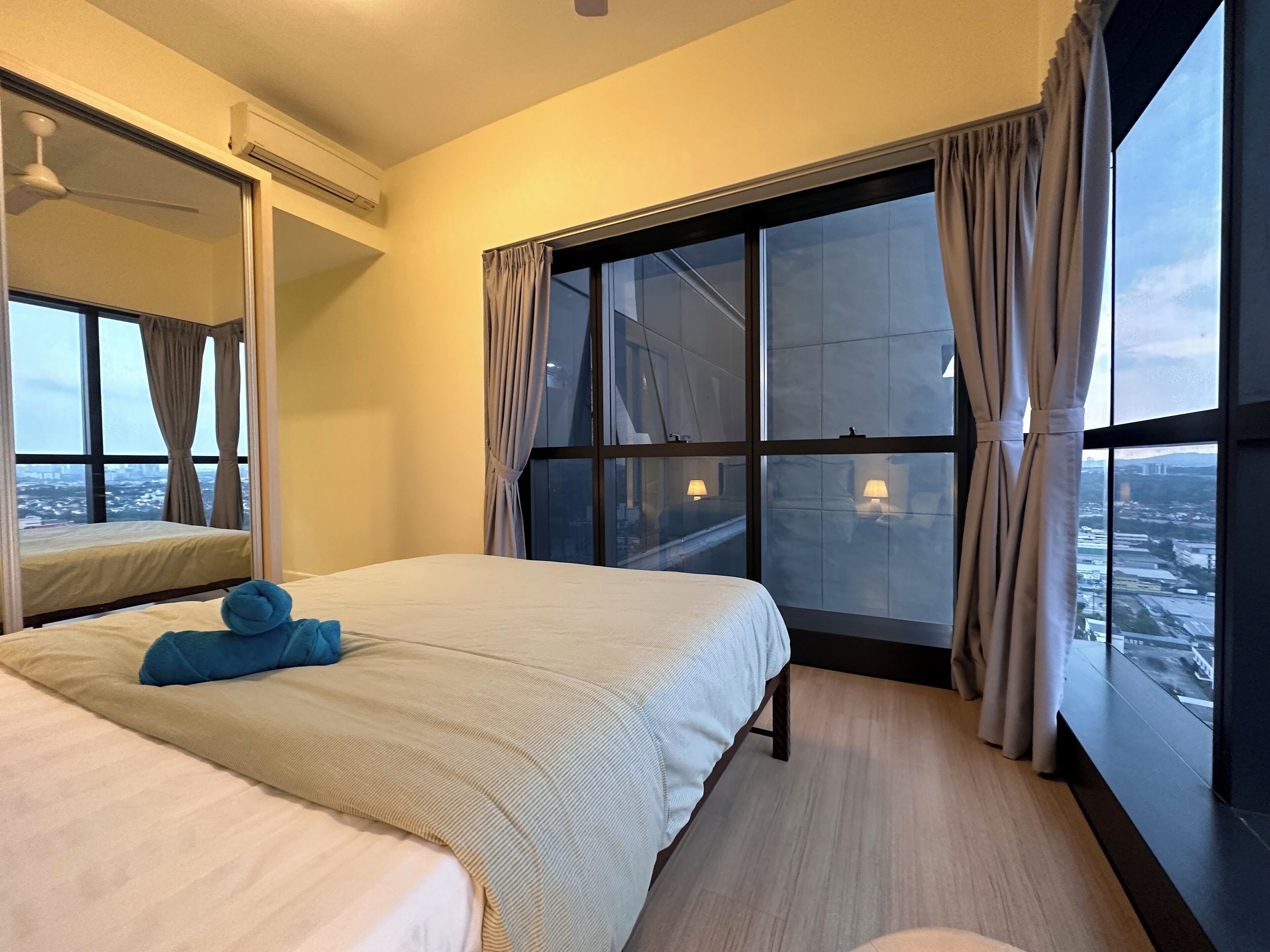 A hotel room with a large bed, a view of the city through floor-to-ceiling windows, and a ceiling fan.