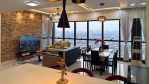 A modern living room with a large TV, a dining area, and a view of a cityscape through large windows. - Skyline Luxe 4BR Retreat with En-suites (Petaling Jaya)