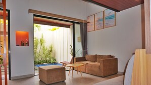 A modern living room with a sofa, ottoman, and a small table. There is a sliding glass door leading to a balcony with plants. - The New Moon Umalas (Kerobokan)