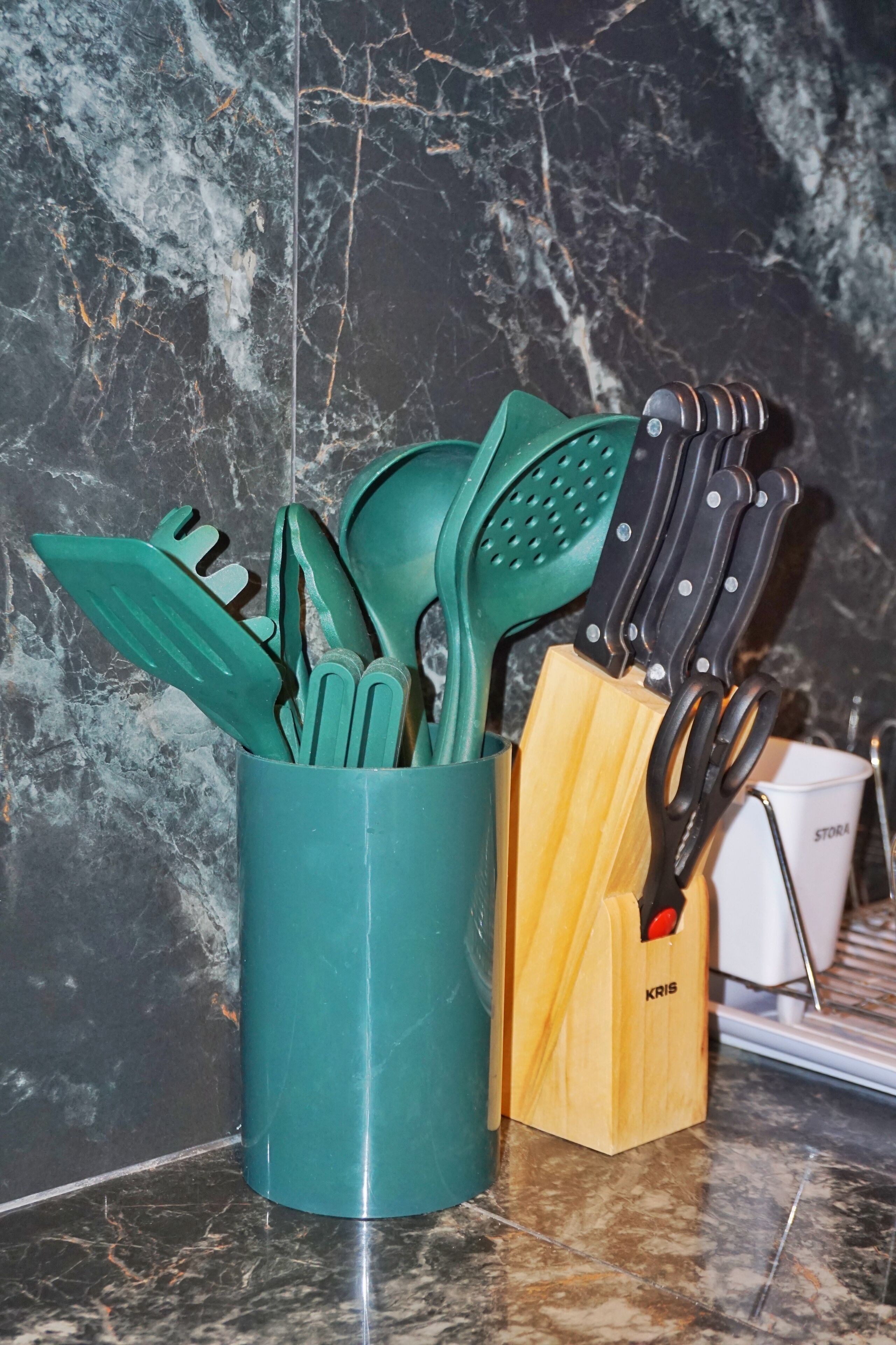 A kitchen utensil holder with green and black utensils, a wooden block, and a pair of scissors on a marble countertop.