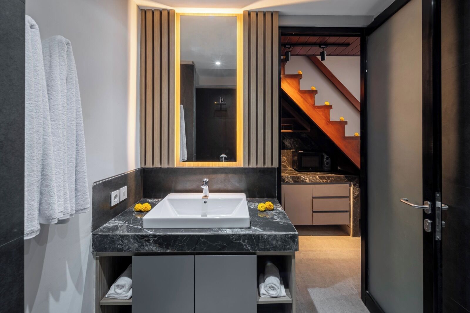 A modern bathroom with a large mirror, a white sink, and a dark marble countertop. There is a towel rack with a white towel and a small yellow flower on the countertop.