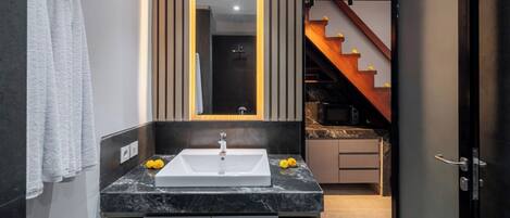 A modern bathroom with a large mirror, a white sink, and a dark marble countertop. There is a towel rack with a white towel and a small yellow flower on the countertop.