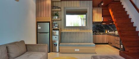 Una cocina moderna con estufa integrada, refrigerador y cafetera. Se ve una escalera de madera que sube a un nivel superior.
