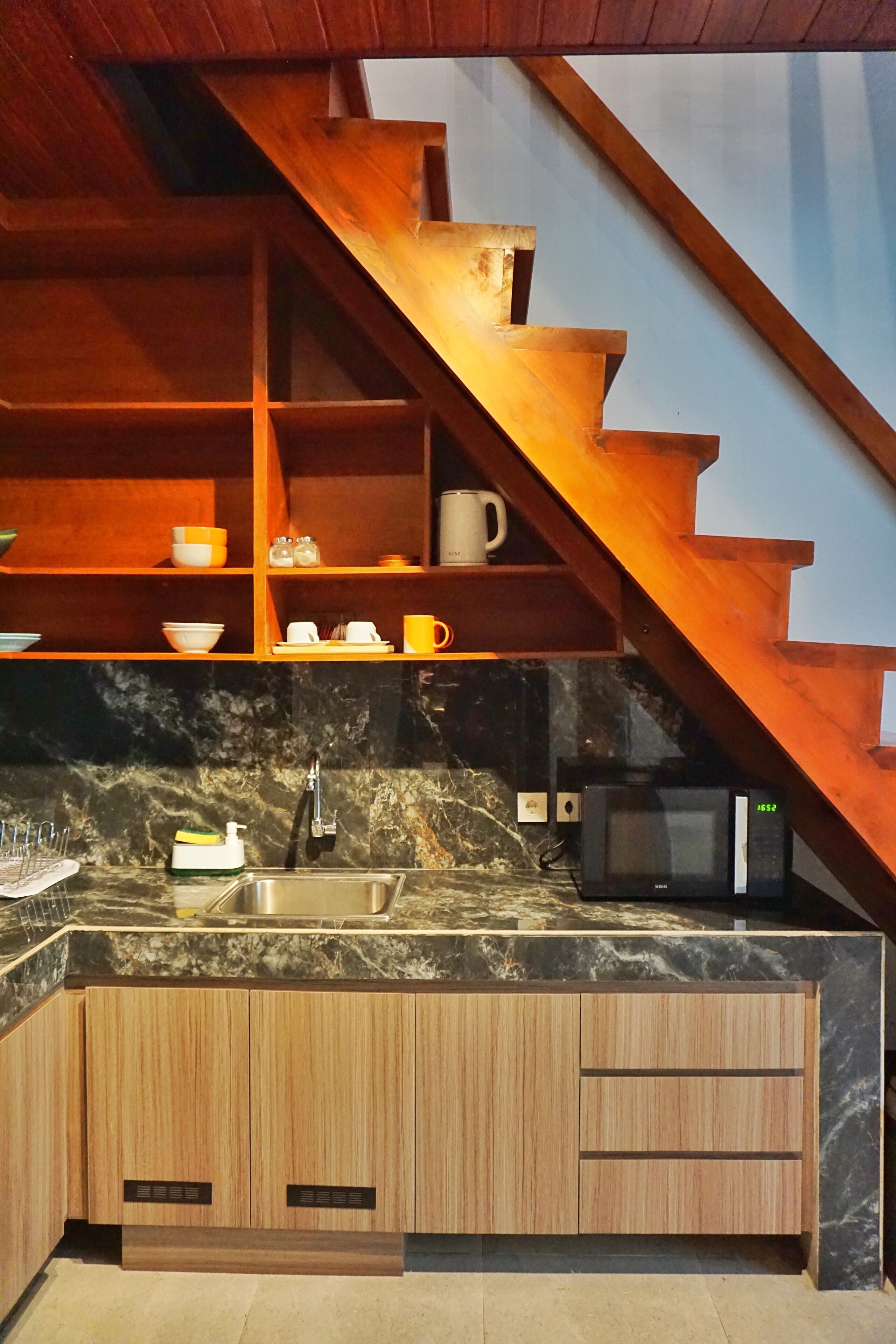 A kitchen with a wooden staircase, a sink, a microwave, and shelves with dishes.