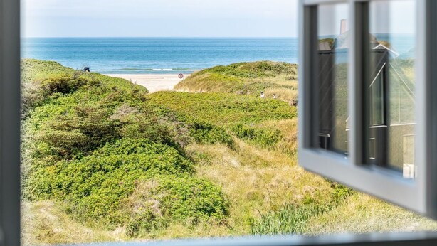 Blick aus einem Fenster auf einen Strand mit Dünen und dem Meer.