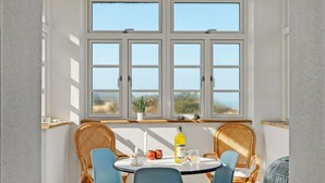 Ein Essbereich mit einem runden Tisch, zwei blauen Stühlen, Korbstühlen, einem Fenster mit Blick und einer Flasche gelber Flüssigkeit auf dem Tisch.