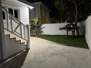 Un quartier résidentiel avec un chemin en béton menant à une maison blanche dotée d’un balcon. On y trouve deux bananiers et une petite pelouse.
