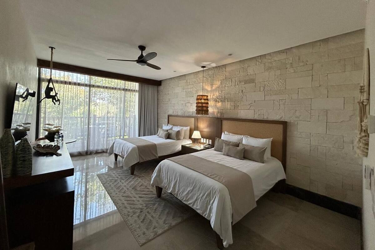 Quarto de hotel moderno com duas camas, uma janela grande com cortinas, uma parede de pedra e um ventilador de teto.