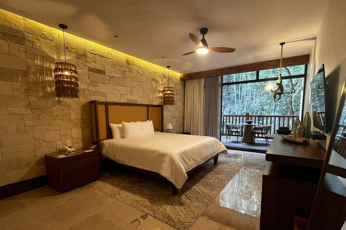 Quarto moderno com parede de pedra, cama grande, escrivaninha e varanda com vista para árvores.