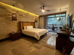 A modern bedroom with a stone wall, a large bed, a desk, and a balcony with a view of trees. - Hotel Mundo Maya Calakmul (Calakmul)