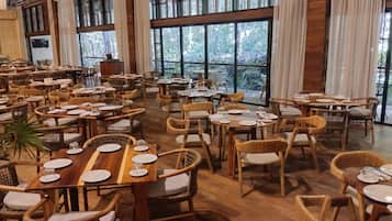 A spacious restaurant with multiple tables and wicker chairs, large windows offering a view of the outdoors, and a wooden floor.