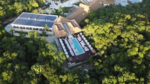Vista aérea de un complejo turístico con piscina, paneles solares y un edificio de techo de paja rodeado de bosque denso.