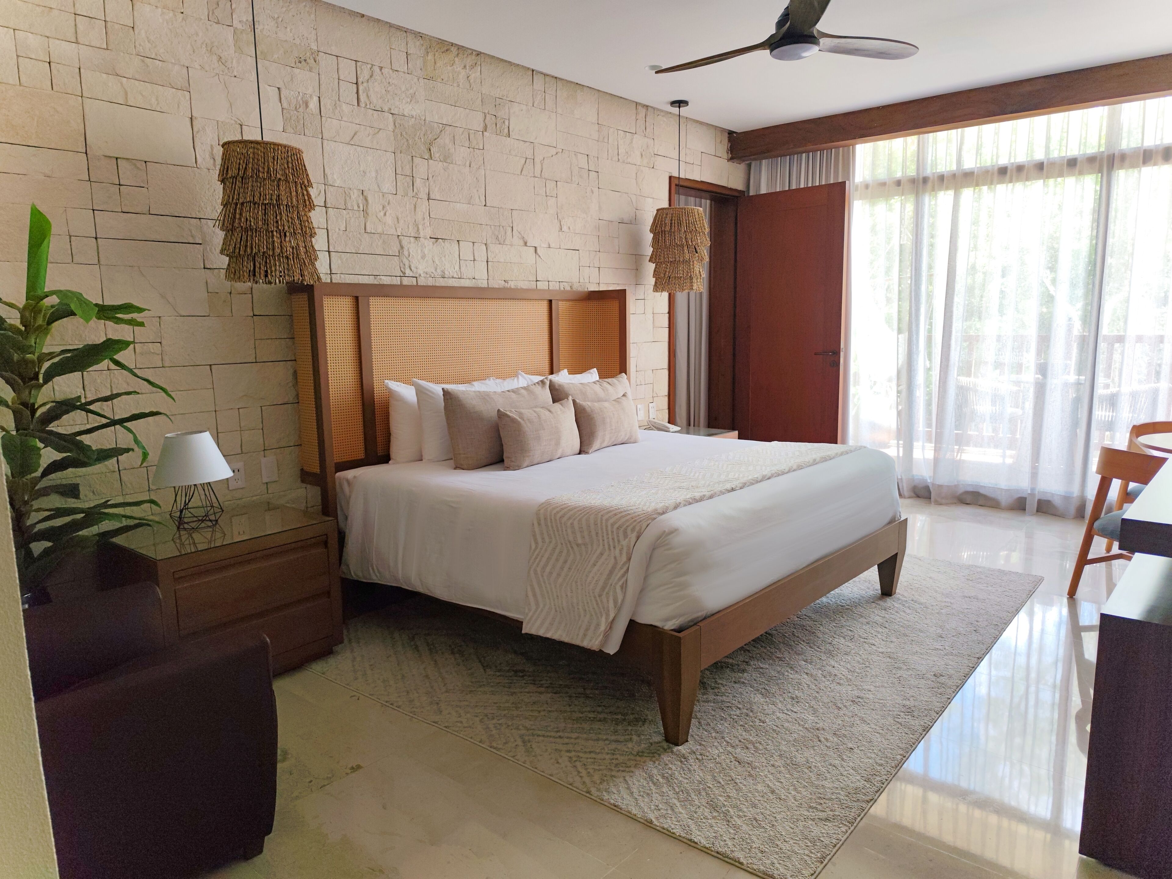 A hotel room with a stone wall, a wooden bed, a nightstand, a lamp, a plant, and a view of the outdoors through large windows.
