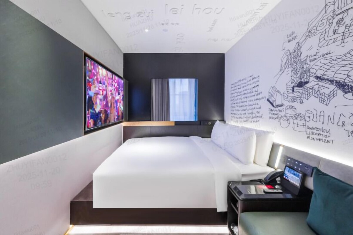 A modern hotel room with a large bed, a desk with a computer, and a wall with architectural drawings.