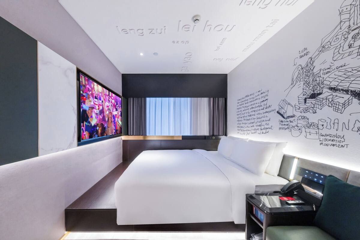 A modern hotel room with a large bed, a flat-screen TV, a desk with a computer, and a wall with sketches and text.