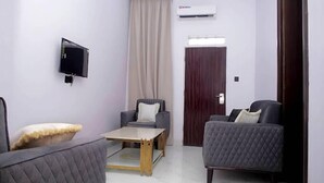 A modern living room with a flat-screen TV mounted on the wall, a sofa, a chair, and a small table. - V City Lodge Hotel and Suites (Abuja)