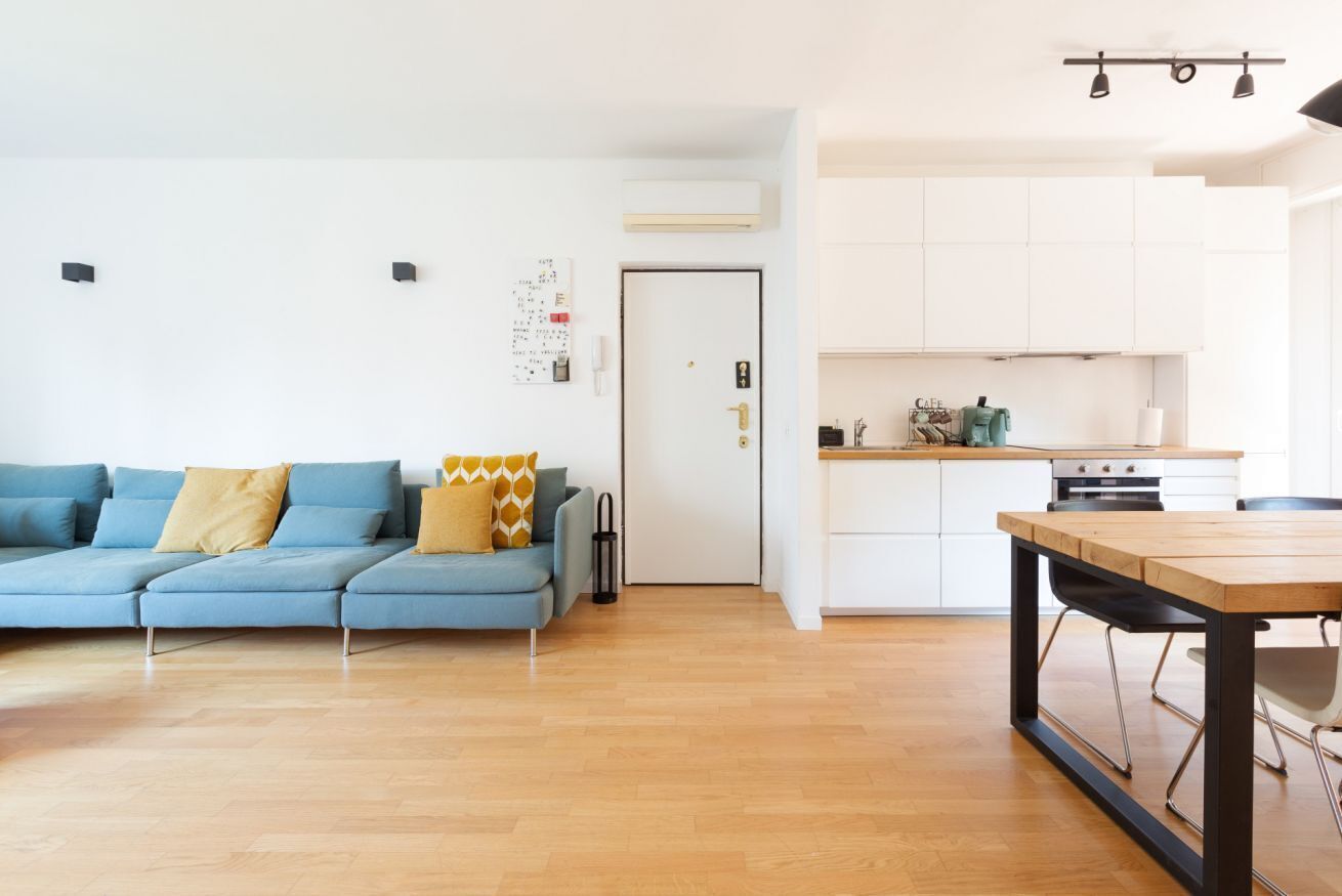 A modern living room with a blue sofa, a wooden dining table, and a kitchen area with white cabinets and a stove.