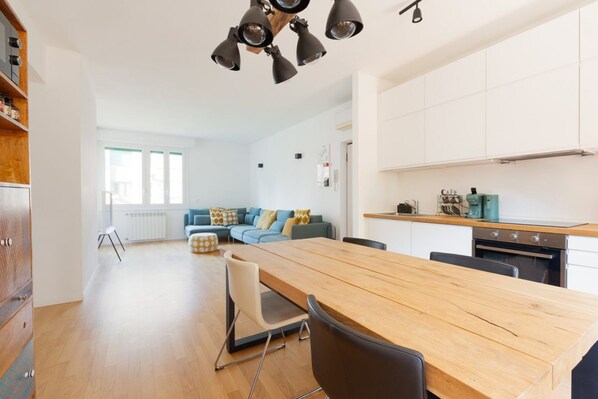 A modern kitchen with a wooden table and chairs, a dining area with a sofa and cushions, and a view of the city through large windows.