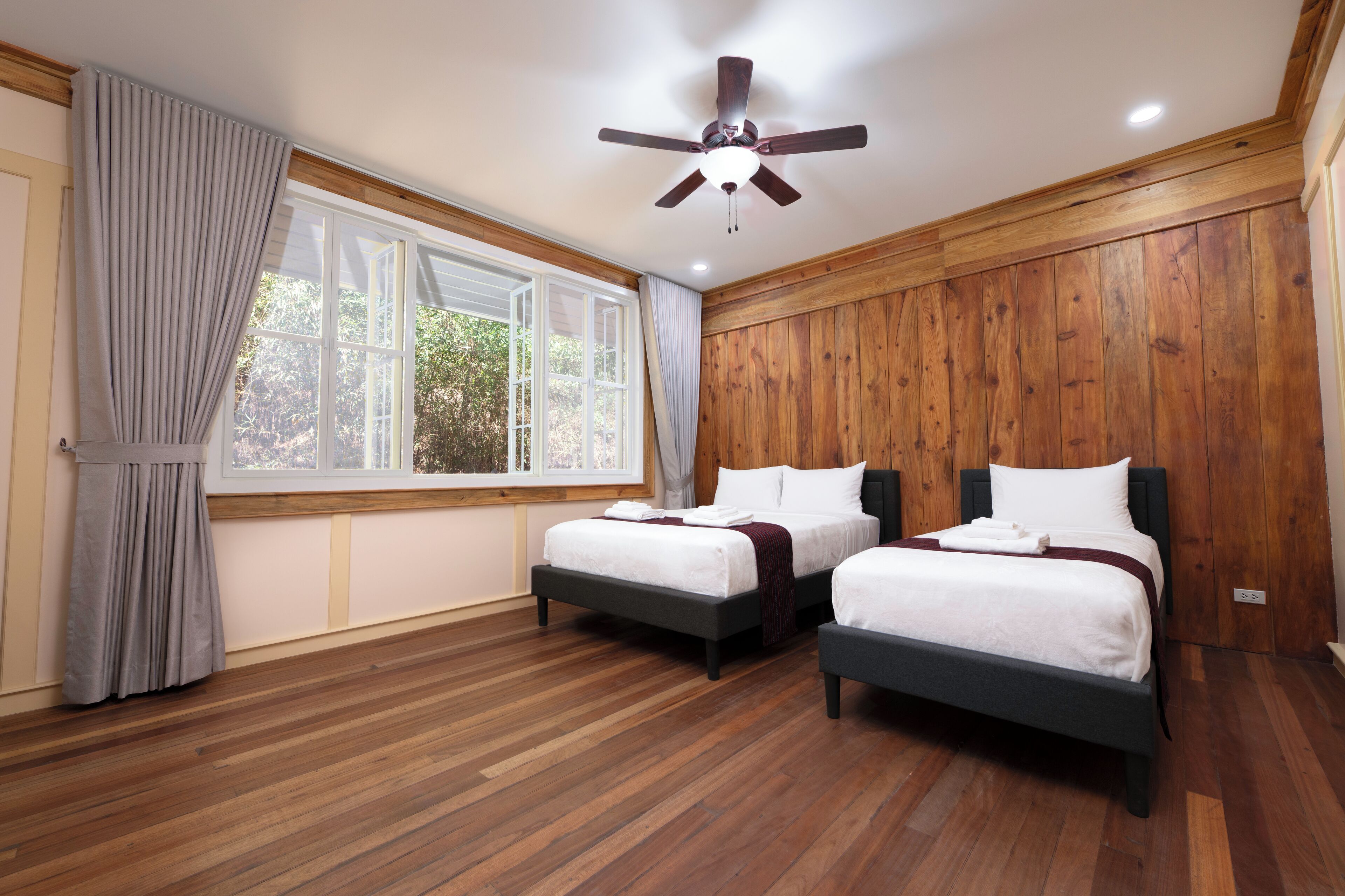 A bedroom with two beds, wooden flooring, a ceiling fan, and a large window with curtains.