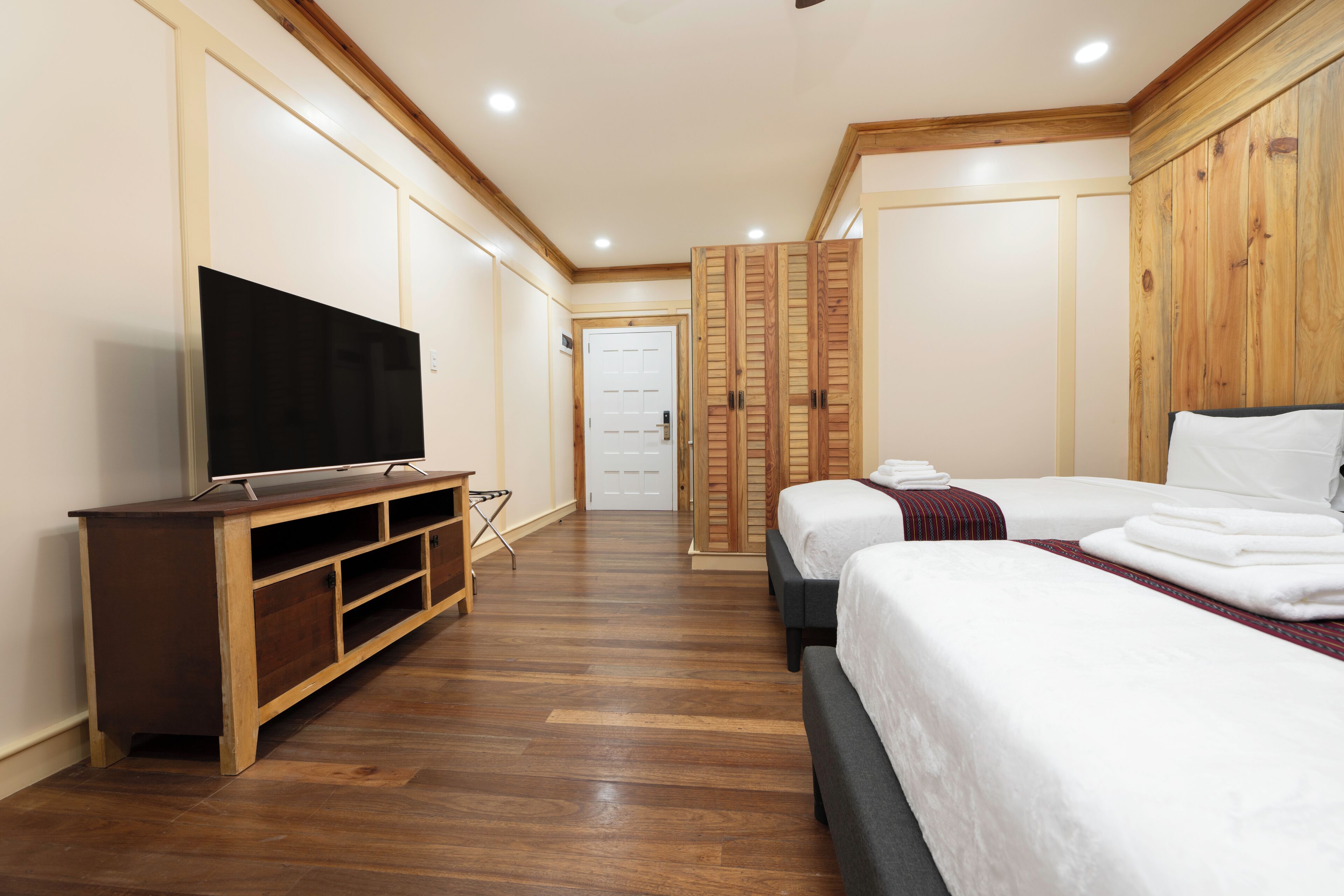 A hotel room with a wooden floor, a TV on a wooden stand, two beds with white bedding, and a wardrobe with wooden doors.