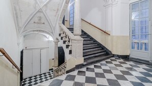 Uma imponente escadaria com balaústres brancos e degraus pretos, que conduz a um ambiente com piso xadrez e uma grande janela.
