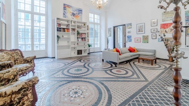 Uma sala de estar ampla com piso estampado, um sofá branco, uma poltrona floral, uma estante de livros e um lustre.