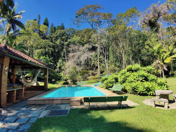 Relax by the serene pool surrounded by lush greenery and trees, with a hammock and seating areas nearby.