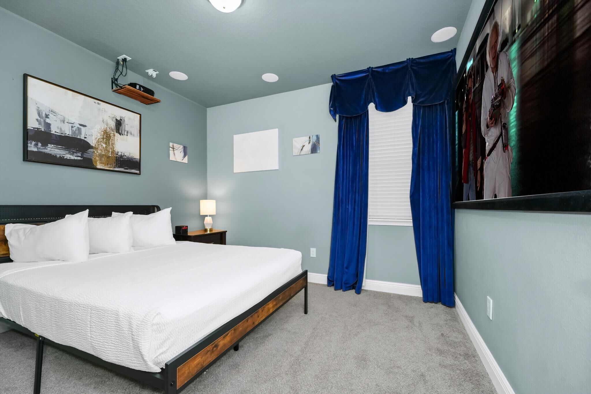 Une chambre avec un grand lit, une table de chevet avec une lampe, un luminaire au plafond et une fenêtre avec des rideaux bleus.