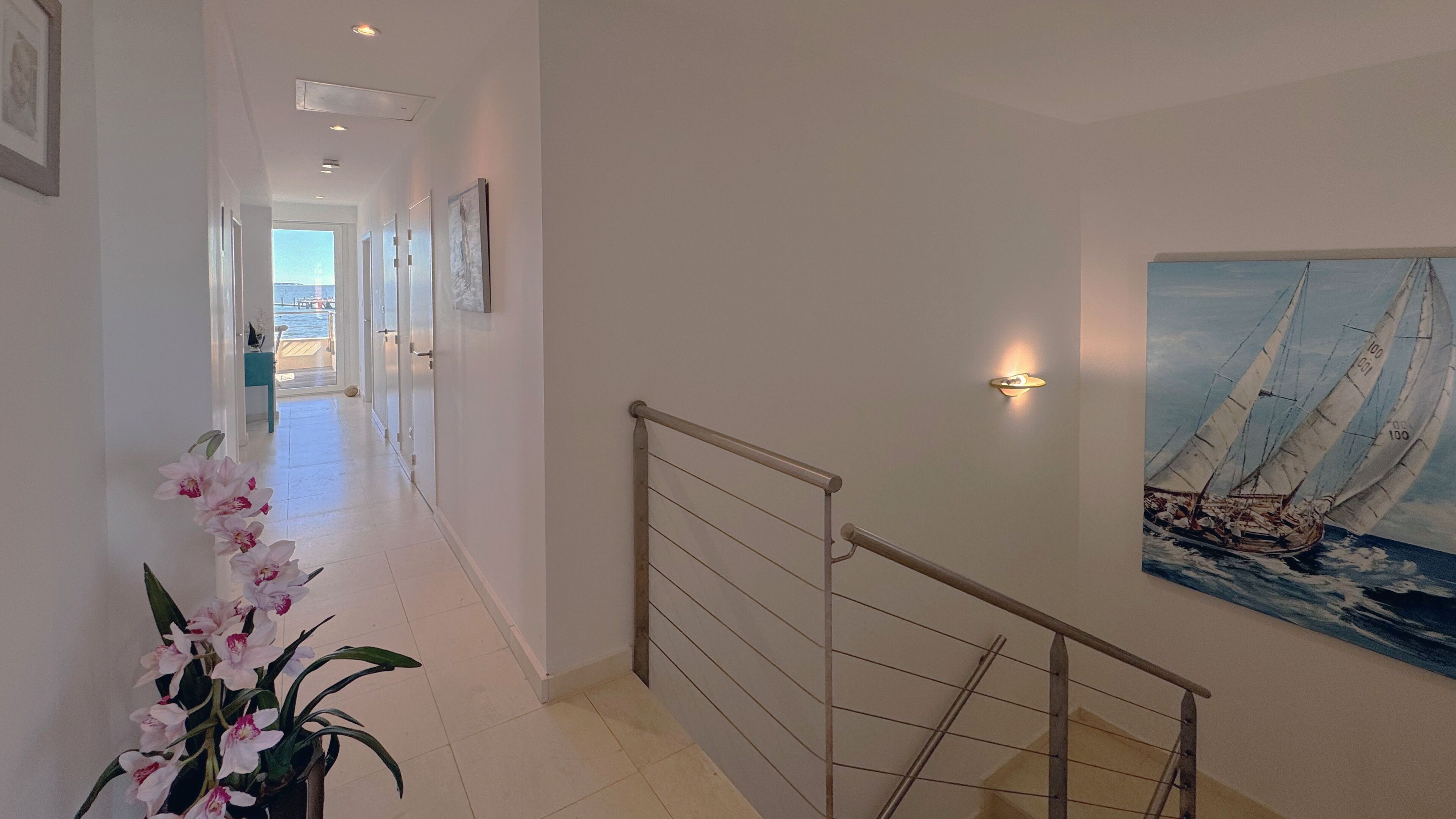 A hallway with a staircase, a potted orchid, and a painting of a sailboat.