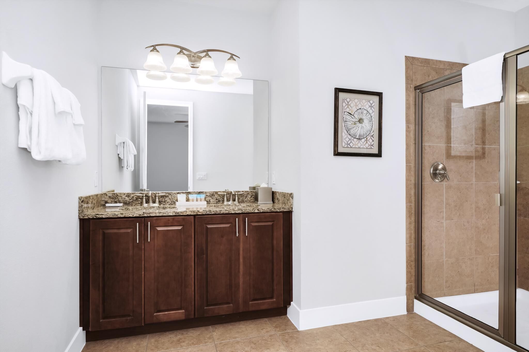 A bathroom with a granite countertop, dark wooden cabinets, a large mirror with lights, a framed artwork, and a glass shower enclosure.