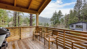 A wooden deck with a grill and chairs, overlooking a forest and a cabin. - Walk to Beach, Arch Cape Getaway w/ Vibrant Interior | Cape Wonder by AvantStay (Arch Cape)