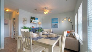 A dining area with a table set for four, chairs, a ceiling fan, and a sofa with a view of the beach.