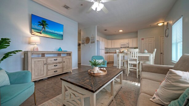 A spacious living area with a large painting of a beach, a ceiling fan, and a dining area with white furniture.