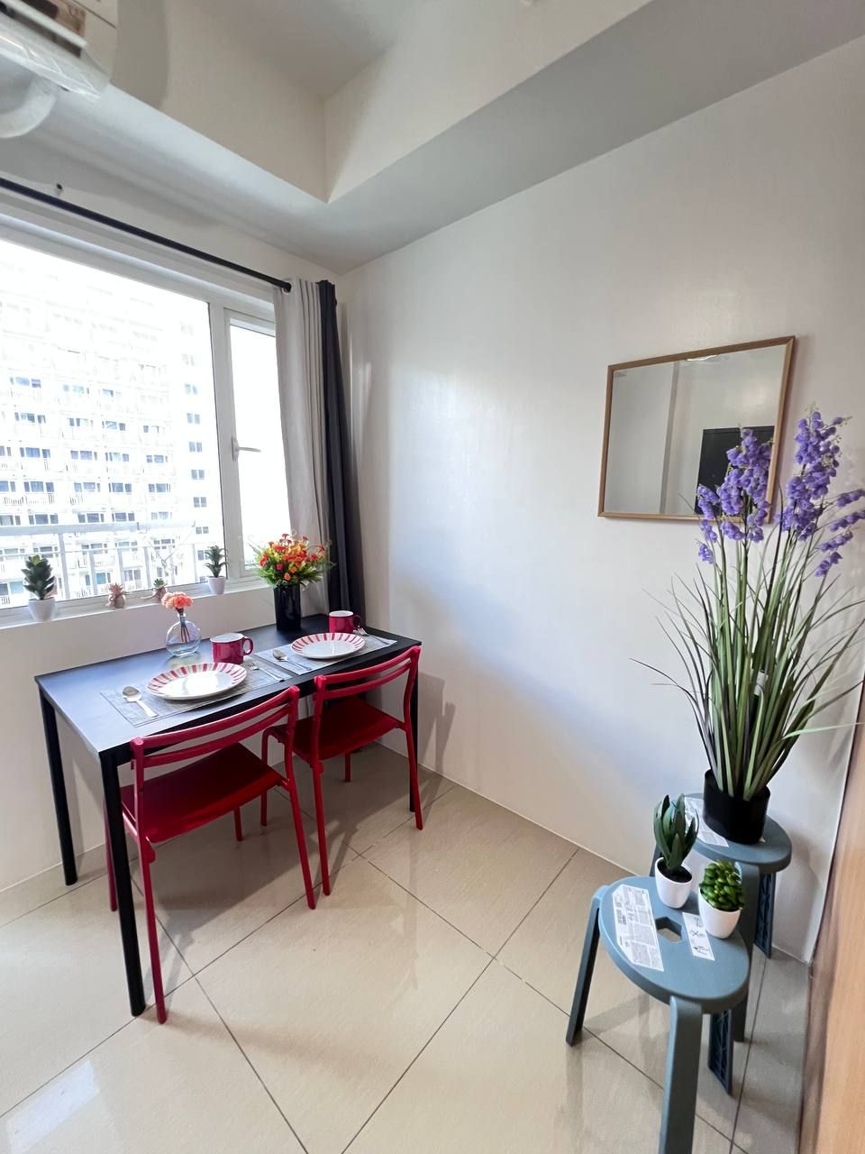 A small dining area with a table and two chairs, a window with curtains, a mirror on the wall, and a potted plant.