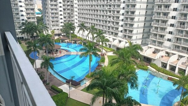 A high-rise hotel with multiple balconies, a central swimming pool, and surrounding palm trees. - 1 BR Shell Residences Facing amenities (Pasay)