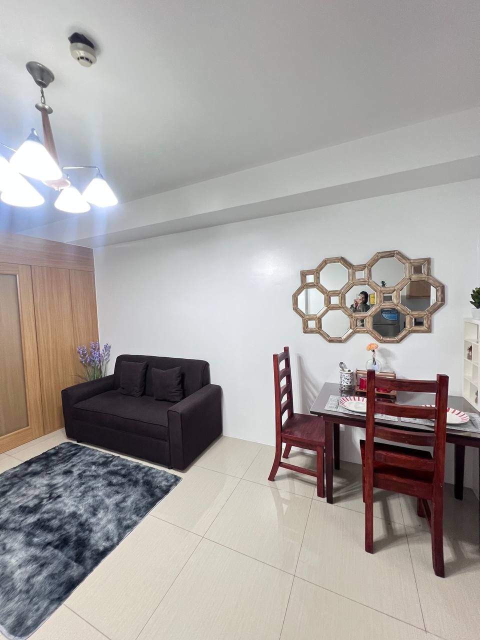Una sala moderna con un sofá oscuro, una mesa de comedor con sillas, un espejo hexagonal y una lámpara de techo.
