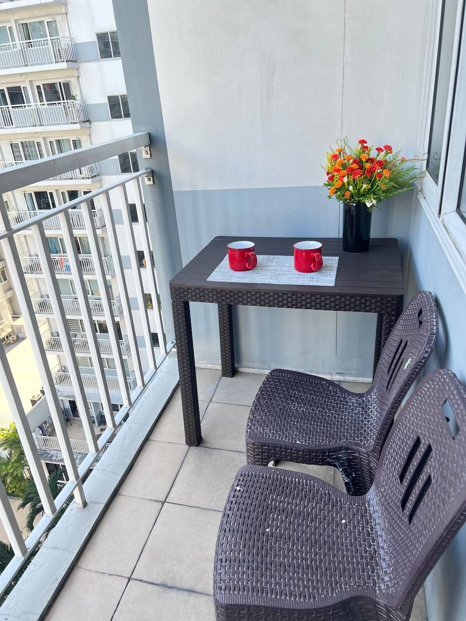 A balcony with a wicker table, two red mugs, and a vase with flowers.