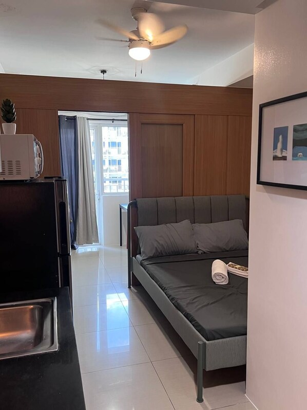 A modern hotel room with a bed, a TV, a ceiling fan, and a view of the city through a window. - 1 BR Shell Residences Facing amenities (Pasay)