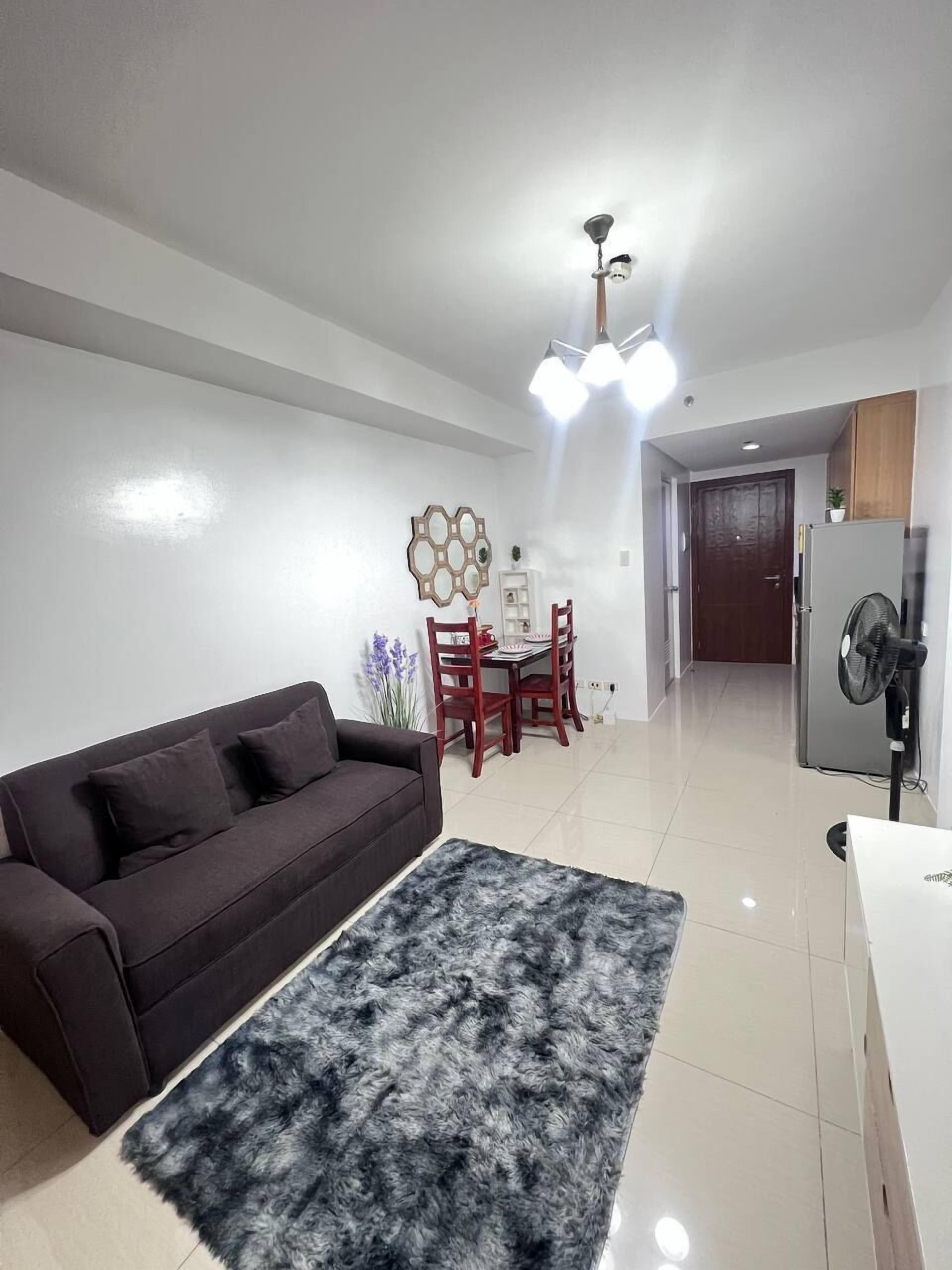 A living room with a sofa, a rug, a dining table with chairs, a ceiling light fixture, and a refrigerator.